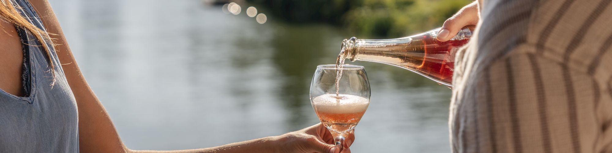 A person is pouring a drink into a glass held by another person, standing on a balcony overlooking a river and trees.