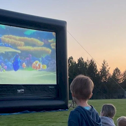 An outdoor movie screening on an inflatable screen at sunset, with children watching from the grass.