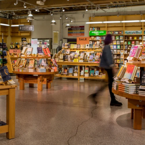 A bookstore interior with shelves full of books and a blurred figure walking. Bright lights illuminate the scene, creating a cozy atmosphere.
