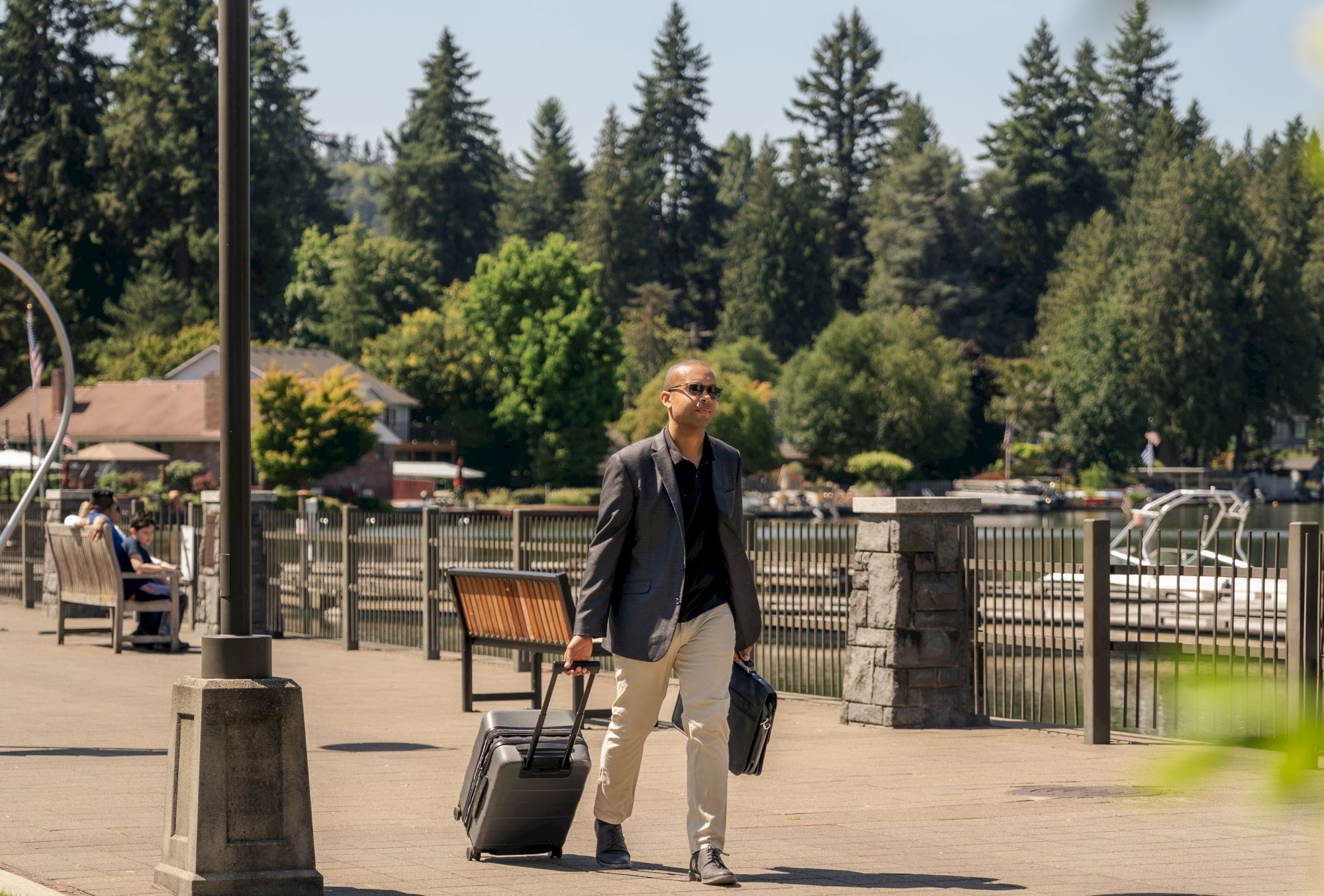 A person is walking along a riverside path with rolling luggage on a sunny day, surrounded by trees and park amenities.