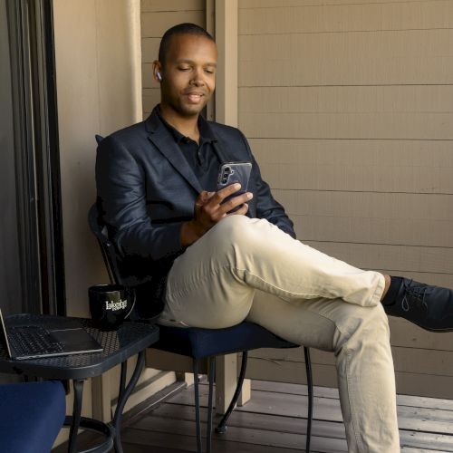 A person is sitting on a chair outdoors, using a smartphone. A laptop and mug are on the table beside them.
