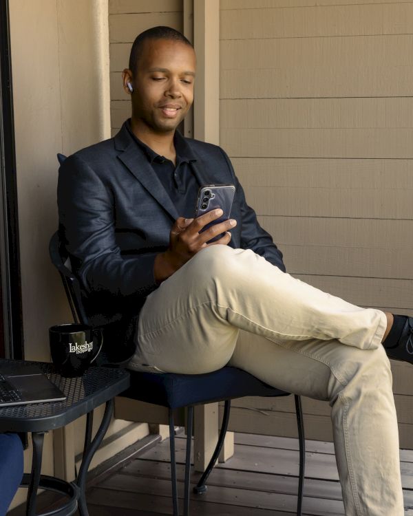 A person is sitting on a chair outdoors, using a smartphone. A laptop and mug are on the table beside them.