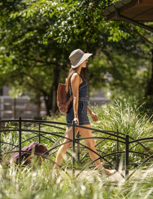 A person in a hat walks across a small bridge surrounded by greenery, carrying a backpack by a stone building.