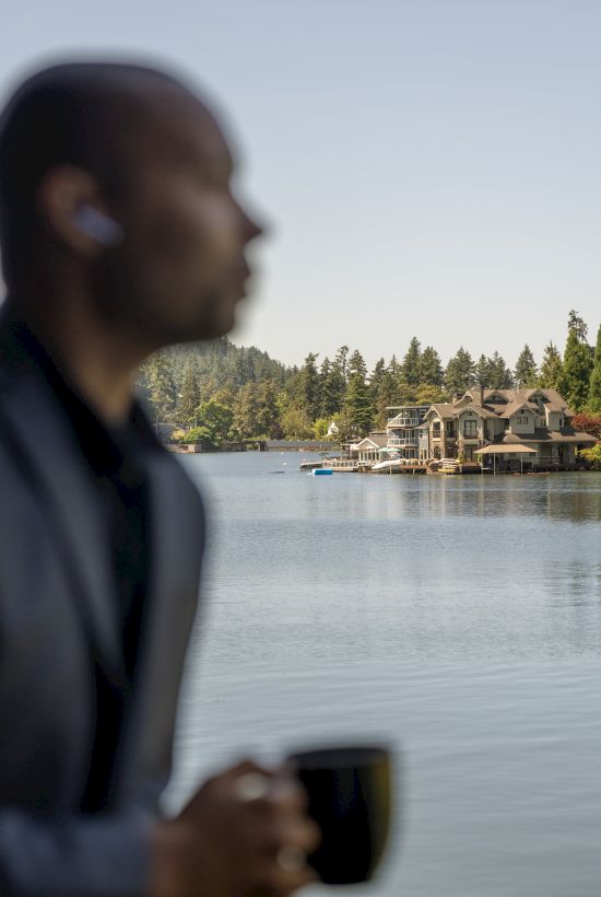 A person holding a mug is blurred in the foreground with a scenic lake and houses in the clear background.