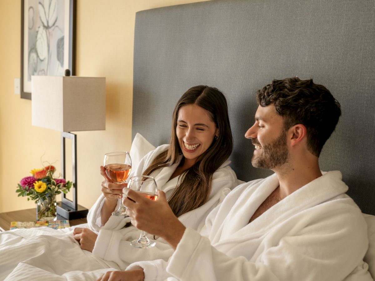A couple in white robes is enjoying drinks in bed, surrounded by a cozy setting with flowers on the side table.