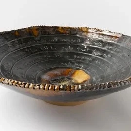 A dark ceramic bowl with a glossy glaze, shallow with a ribbed pattern, some brownish edges, and a small rock or crumb inside the center.