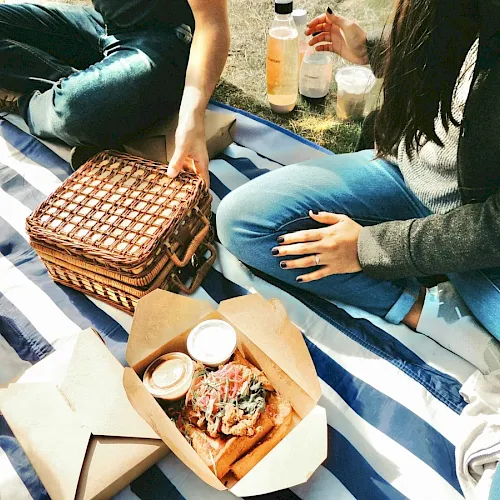 People having a picnic on a striped blanket with food boxes, drinks, and a woven basket.