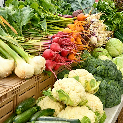 A vibrant display of fresh vegetables: carrots, radishes, cauliflower, broccoli, cabbage, onions, leeks, and greens arranged at a market.