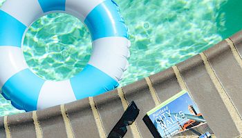 A bright poolside scene with a blue-and-white lifebuoy, a striped towel, sandals, a skateboard, a flip-flop, a can, and a few magazines by the edge of the pool.