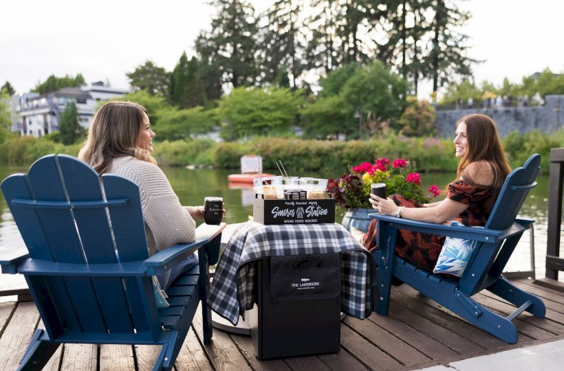 Two women chat on blue Adirondack chairs by a lakeside patio, with coffee and snacks on a cart during a sunny day.