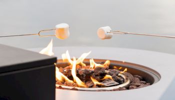 Marshmallows roasting on skewers over an open fire in a backyard grill on a sunny day.
