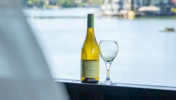 A wine bottle and a glass on a railing by the water, with blurred trees and houses in the background.