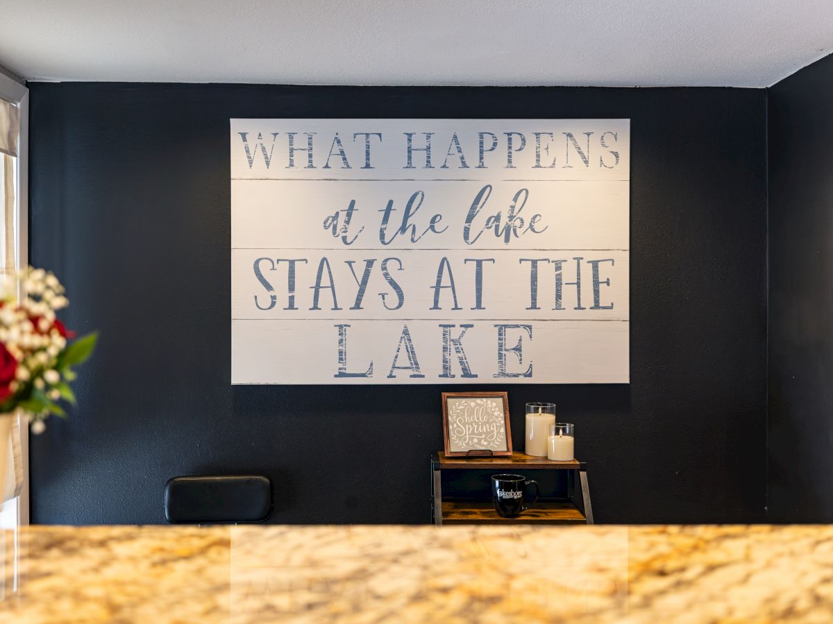 A wall sign reads &ldquo;What happens at the lake stays at the lake,&rdquo; above candles and a &ldquo;Hello Spring&rdquo; card on a small shelf.