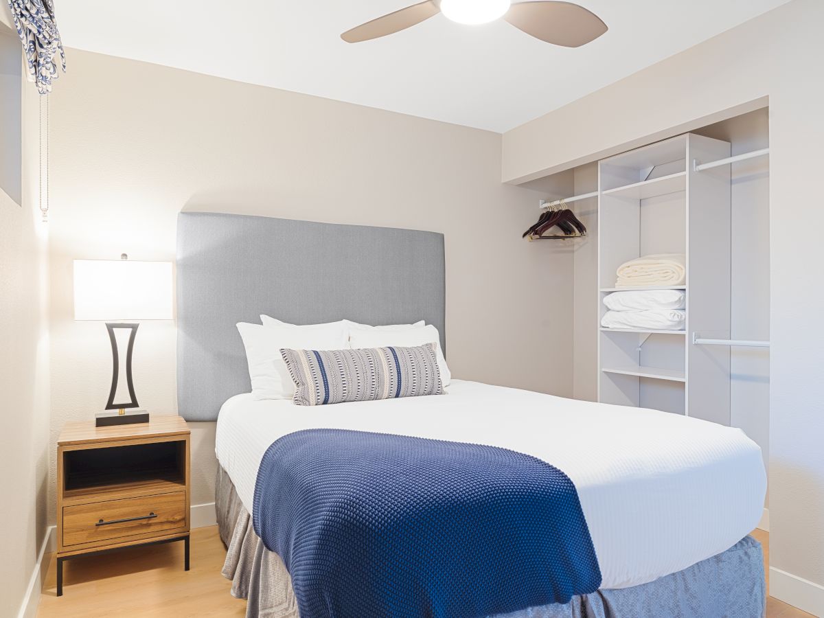 A bright modern bedroom with a bed, blue throw blanket, bedside lamp, ceiling fan, and built-in closet shelves with hangers.