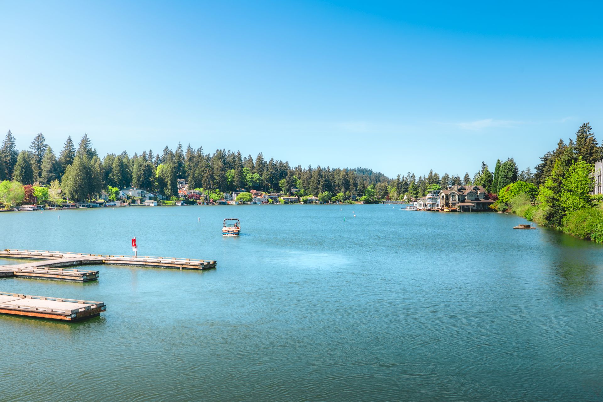 A calm blue lake with docks in the foreground, a small boat in the water, and lakeside houses backed by tall pine trees.