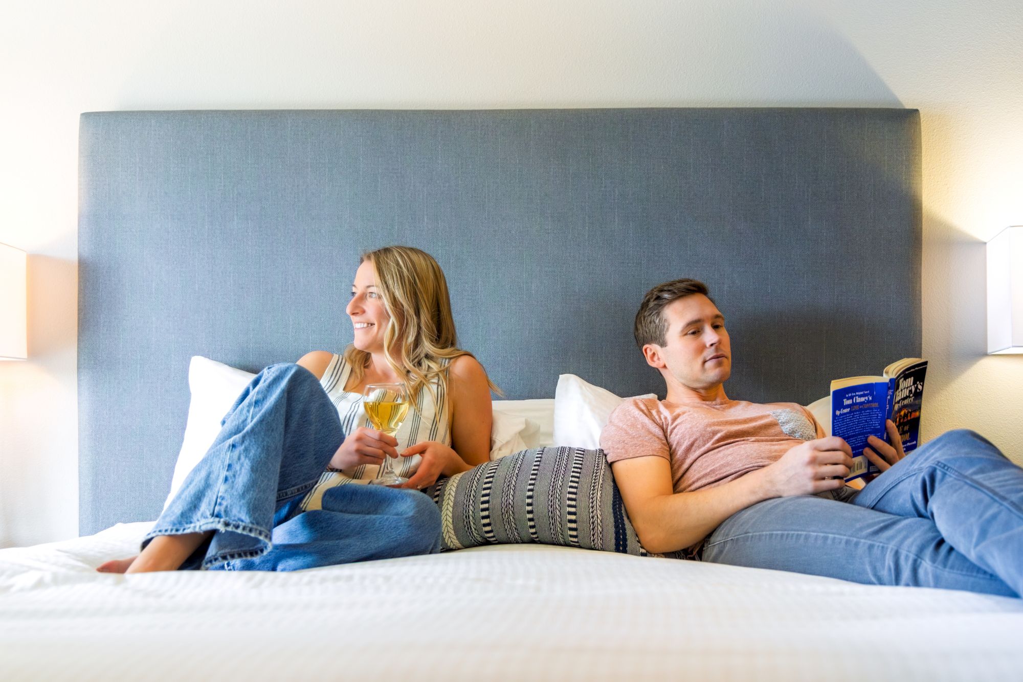 Two people sit on a bed relaxing&mdash;one woman with a drink, one man reading a book&mdash;lifestyle moment in a cozy hotel setting.