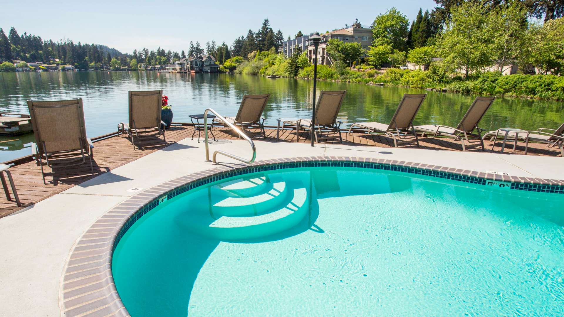 A pool with clear blue water is surrounded by lounge chairs, set against a serene lake and tree-lined landscape.