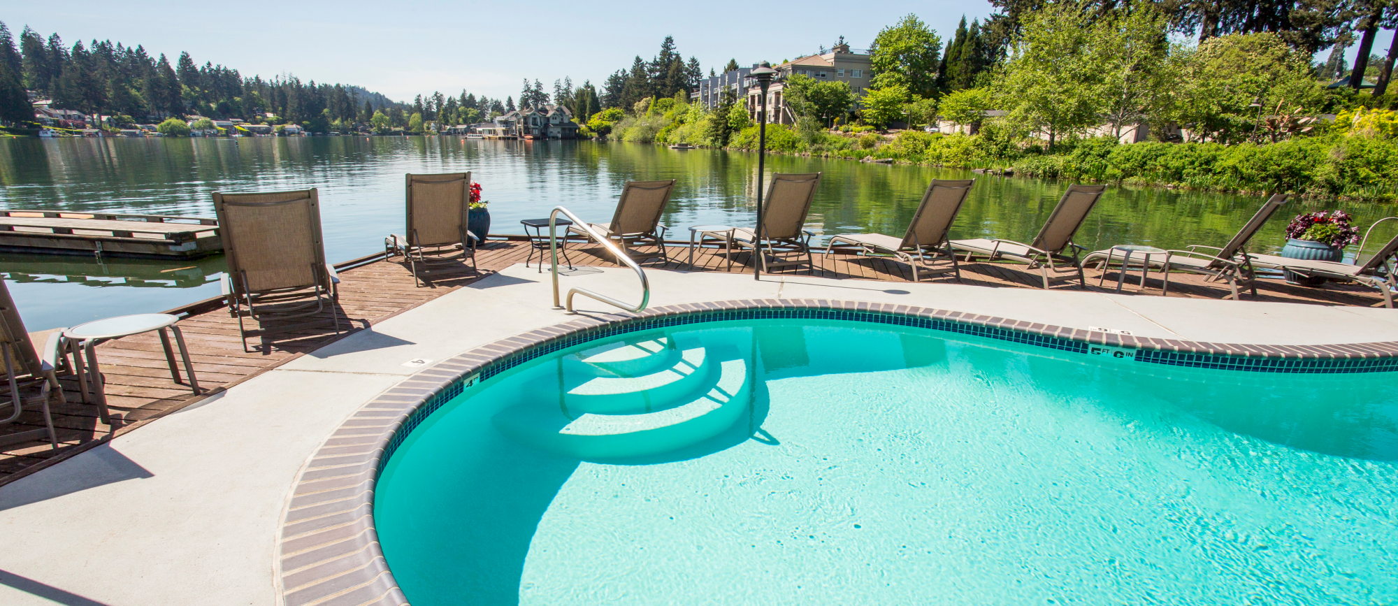 A pool with clear blue water is surrounded by lounge chairs, set against a serene lake and tree-lined landscape.