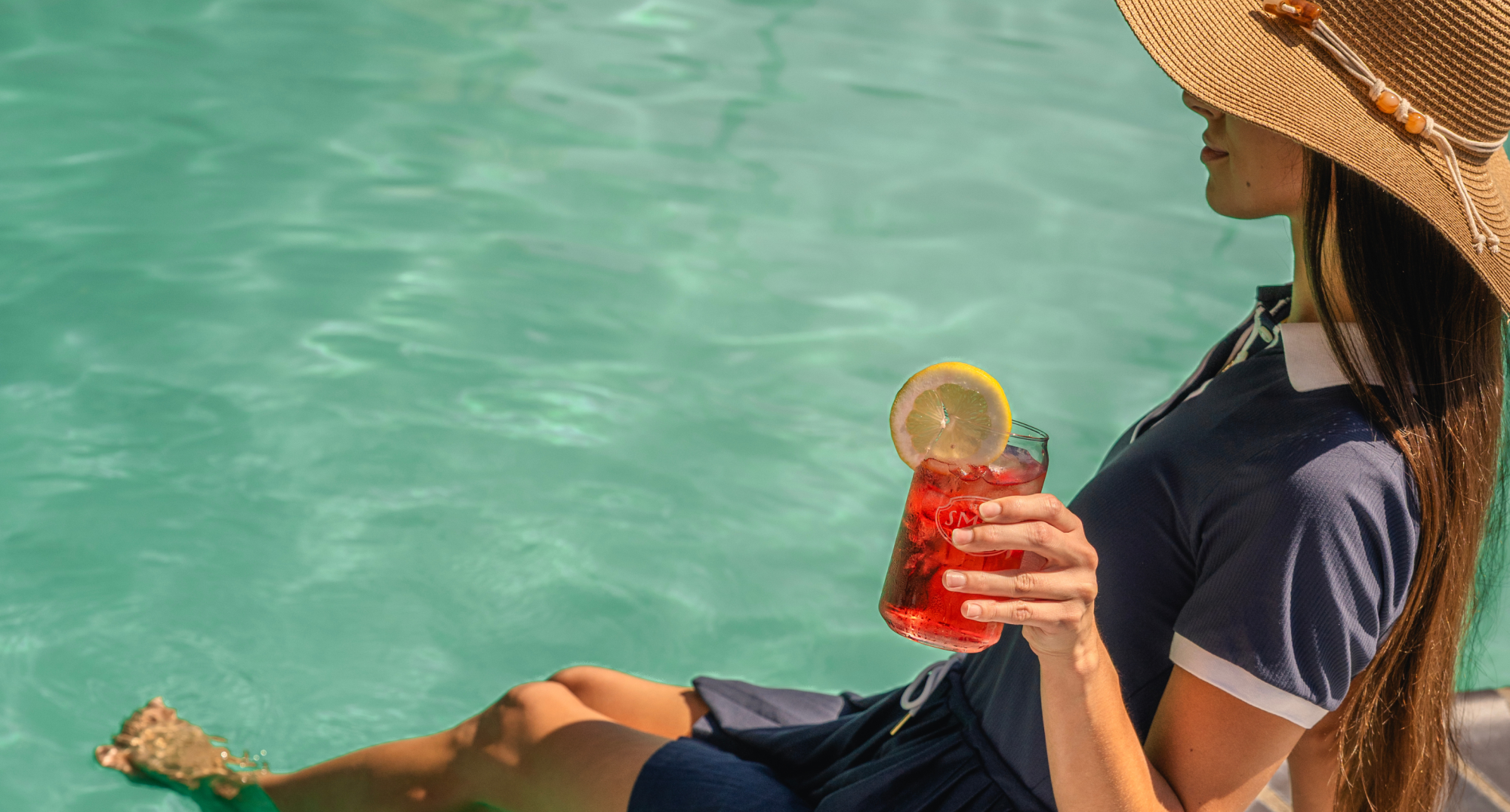 A person in a hat relaxes by a poolside, dipping their feet in the water, holding a drink garnished with a lemon slice.