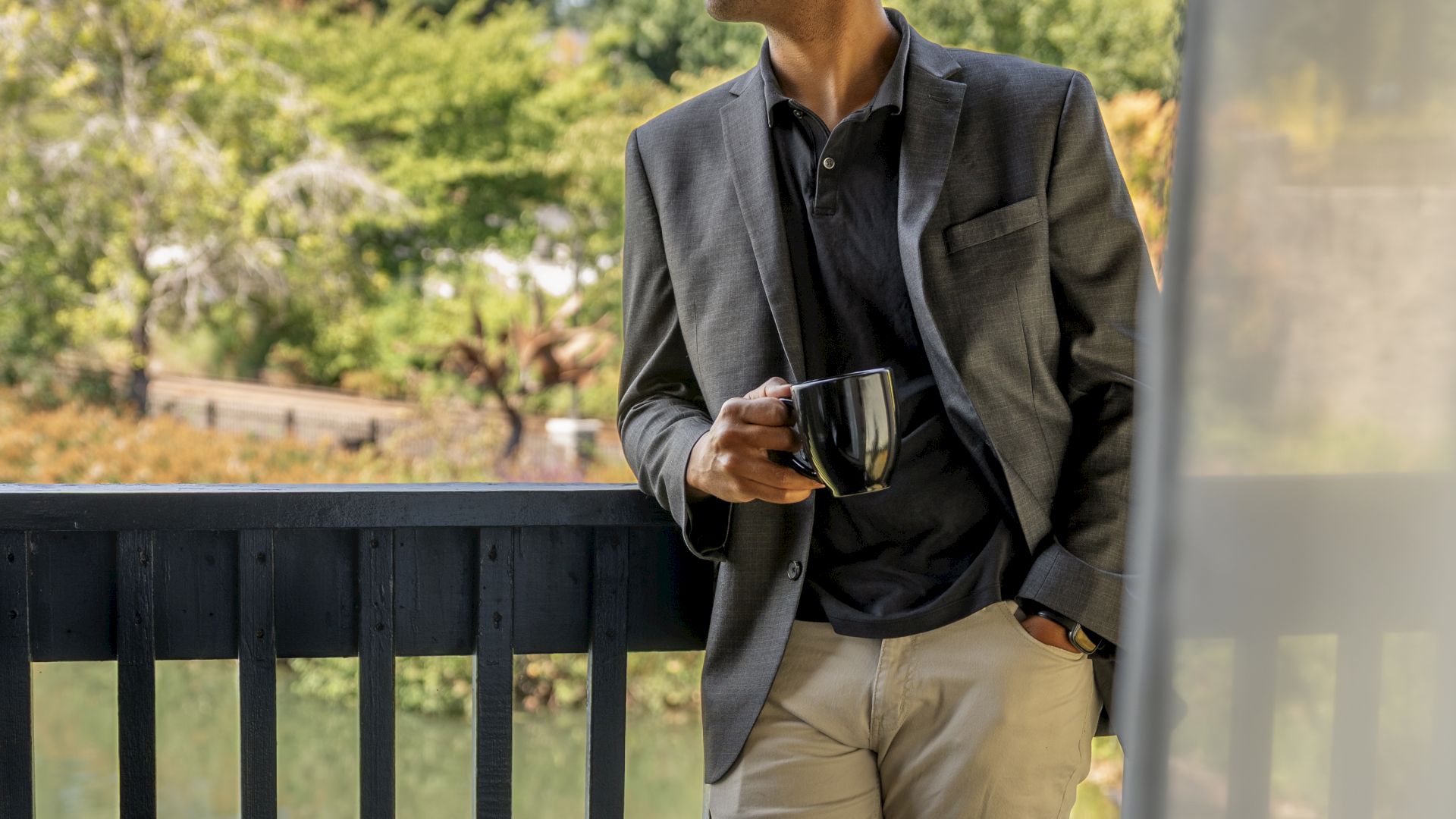 A person in a suit stands on a balcony holding a cup, looking out at a view with trees in the background.