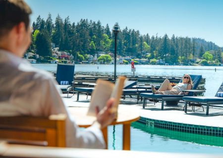 Two people lounge by a pool overlooking a lake with trees; one reads a book while the other relaxes on a chair under clear skies.