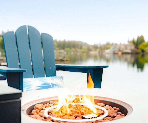 A sunny outdoor scene with a plate of food in the foreground and teal Adirondack chairs by a table, overlooking a beach or waterfront.