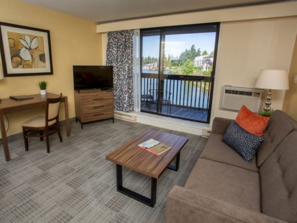 A cozy living room with a sofa, coffee table, TV, desk, and balcony view. Decor includes a painting, lamps, and patterned curtains.