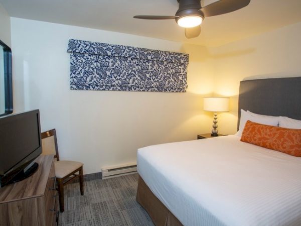 A hotel room with a bed, chair, TV, lamp, and overhead fan. The decor includes a patterned wall hanging and orange accent pillow.