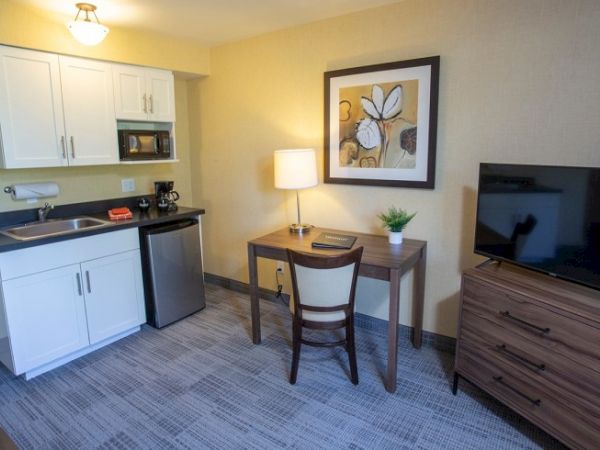 A small room with a kitchenette, table, chair, lamp, TV on a dresser, and a framed floral artwork on the wall.