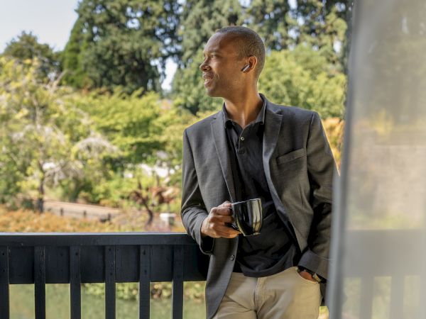 A person wearing a blazer stands on a balcony holding a mug, looking out at a scenic view with trees in the background.