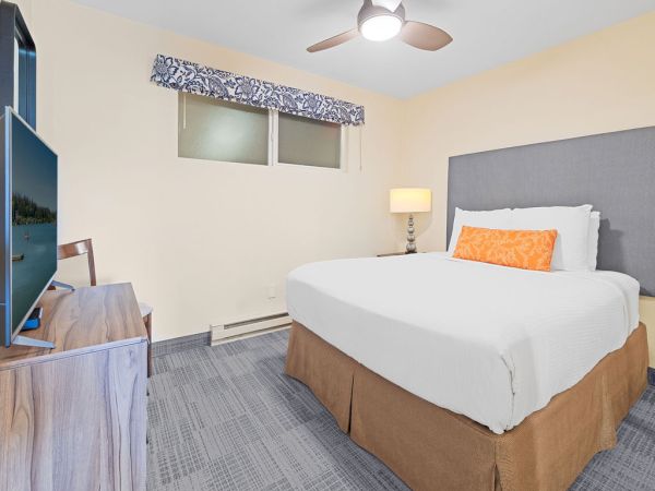 A simple, tidy hotel room with a queen bed, gray headboard, orange accent pillow, ceiling fan, lamp, window, and desk setup.