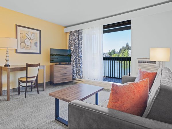 A bright hotel living room with a gray sofa, orange cushions, a wooden coffee table, desk and chair, a TV, yellow wall, and balcony outside.