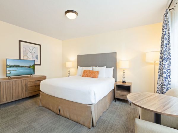 A clean hotel room with a king bed, beige tones, two nightstands, a dresser with a TV, and a small round table by windows with patterned curtains.