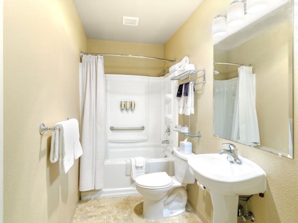 A small, beige-tiled bathroom with a white tub/shower, a white toilet, a pedestal sink, a large mirror, towel bars, and a few shelved toiletries.