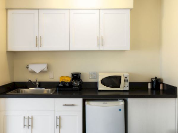 A compact kitchen with white cabinets, black countertop, a sink, coffee maker, microwave, small fridge, toaster, and a blender on the counter.