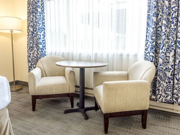 A small sitting area with two beige upholstered chairs around a round table, a tall floor lamp, and blue patterned curtains by a window.