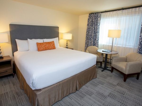 A hotel room with a large bed, two lamps, a small table, and chairs near a window with patterned curtains.