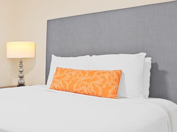A neatly made hotel-style bed with white linens, a gray upholstered headboard, and an orange accent pillow beside a small bedside lamp, cozy and modern.