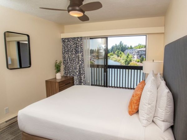 A bedroom with a bed, orange pillow, and window with a view of a lake and trees, features a ceiling fan and wall mirror.
