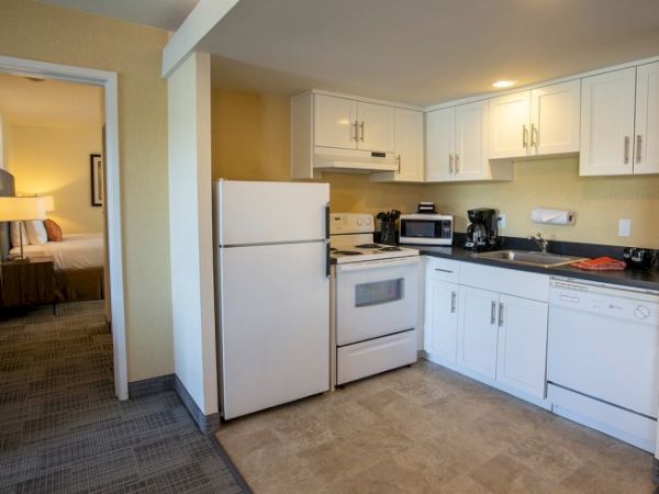 A compact kitchen with white cabinets, a fridge, stove, microwave, and coffee maker, adjacent to a bedroom.