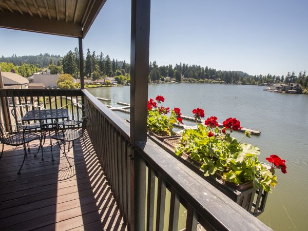 A lakeside balcony with a wooden deck, red geraniums in window boxes, a railing, and a view of boats and trees across the water, sunny day.