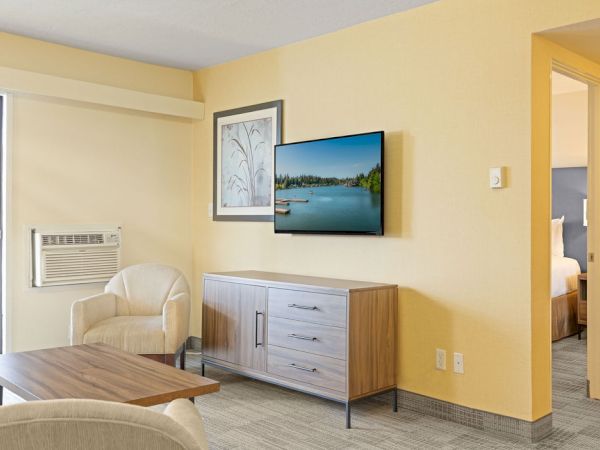A modern hotel living area with a TV on a yellow wall, a wooden cabinet, a framed picture, a beige chair, and a doorway to a bedroom.