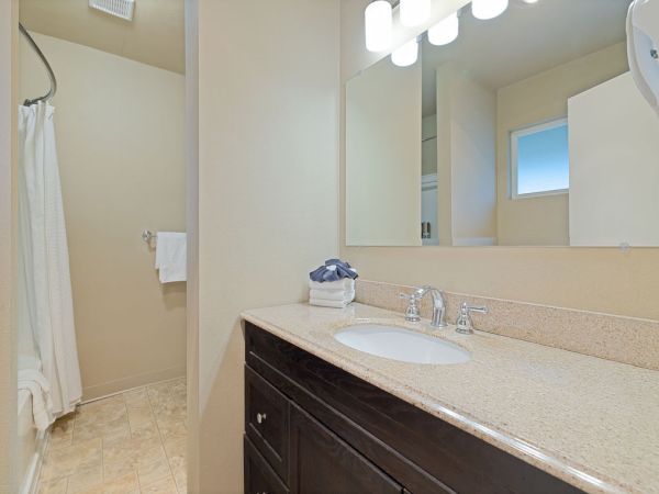 A clean bathroom with a speckled countertop, dark vanity, single sink, mirror, bright overhead lights, a shower curtain, and beige walls.