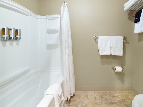 A white bathtub with a shower curtain, wall hooks with toiletries, a towel rack with towels, and a toilet paper holder on a beige tiled bathroom.