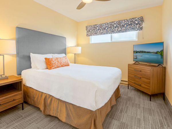 A cozy hotel room with a queen bed, beige walls, blue headboard, two bedside lamps, a window valance, and a wooden dresser with a TV.