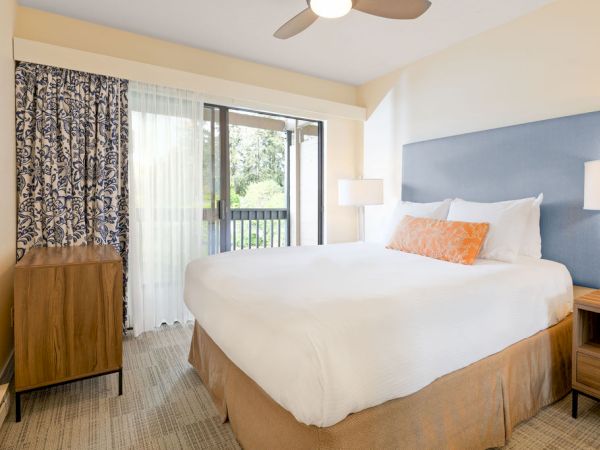 A bright hotel room with a king bed, white bedding, blue headboard, two nightstands with lamps, a wooden dresser, patterned curtains, and a balcony sliding door.