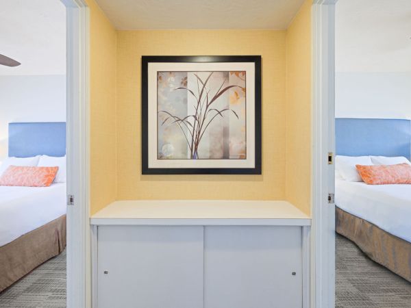 A small built-in display nook with a framed botanical print, pale yellow walls, and a white shelf cabinet between two hotel rooms with twin beds.