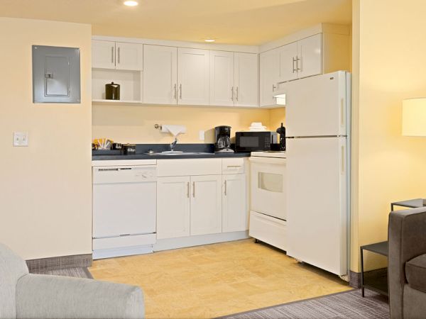 A compact, tidy kitchenette with white cabinets, a white stove and fridge, a small counter area, and a living room in the foreground, bright lighting.