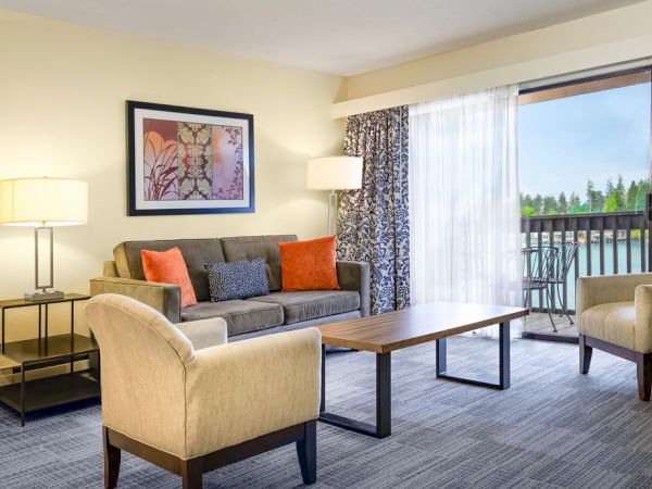 A modern hotel living room with a gray sofa, two beige armchairs, a wooden coffee table, lamps, framed art, and a balcony view through large glass doors.