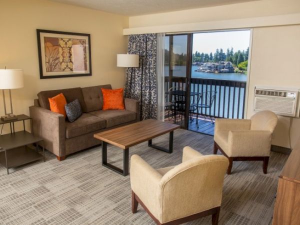 A cozy living room with a sofa, chairs, a coffee table, and a balcony view of a lake. There's a painting on the wall and two lamps.